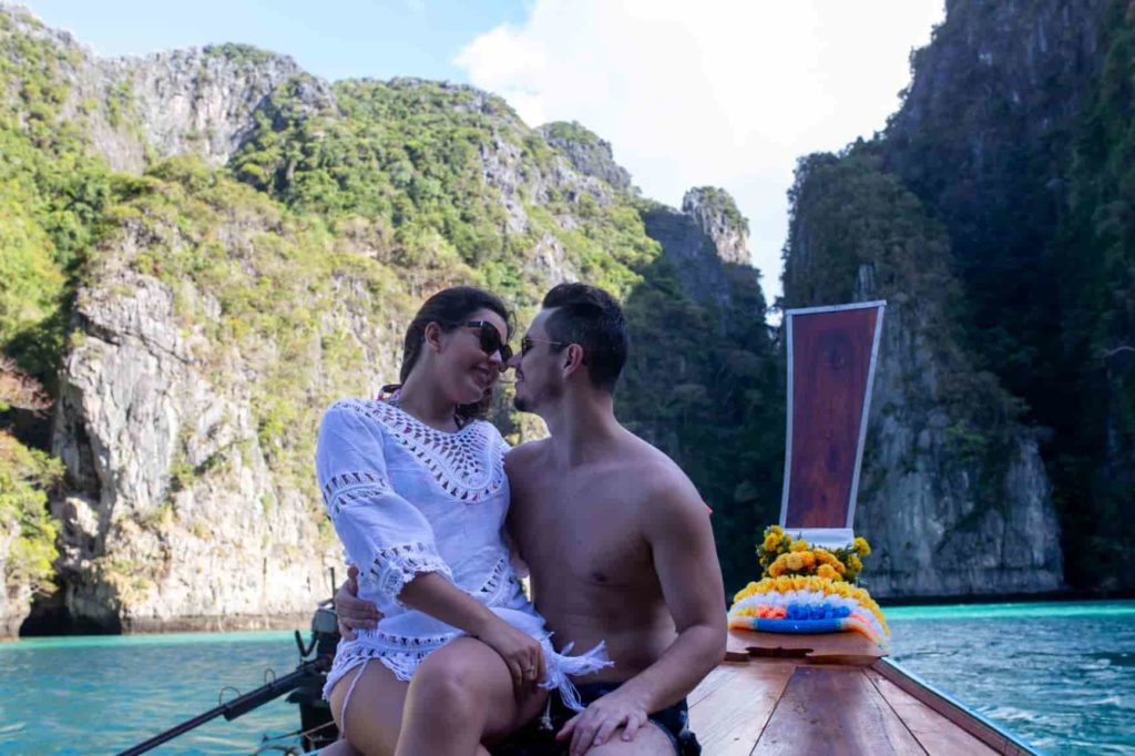 Casal sentado na frente de um barco tailandês, virados de costas, com phileh lagoon no fundo