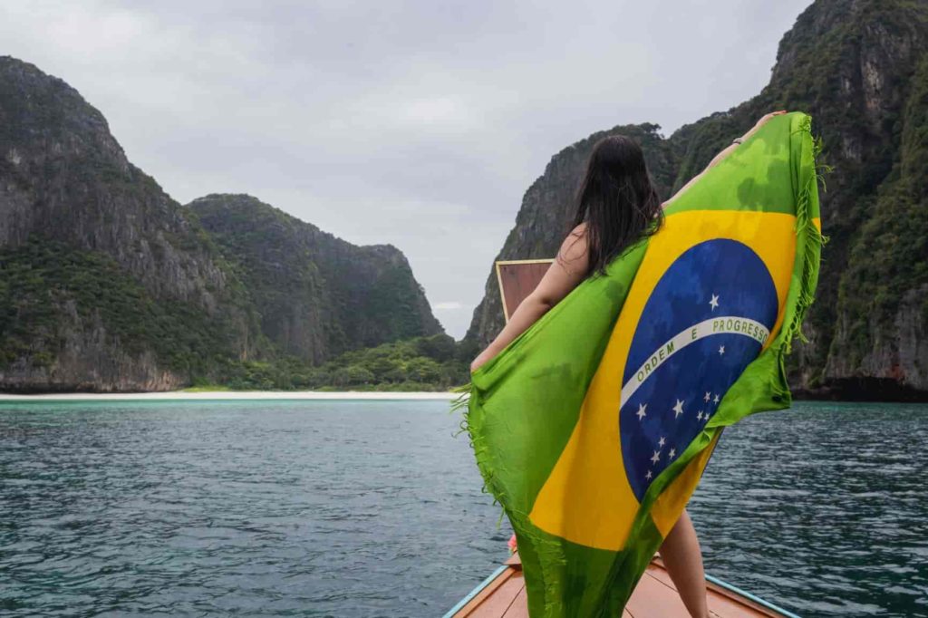 Brasileira em pé na frente de um barco tailandês estendendo a bandeira do brasil em Maya Bay na Tailândia