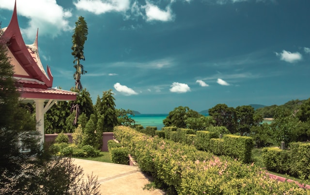 Vista de um jardim bem cuidado com arbustos verdes e uma construção tradicional tailandesa, com o mar e um céu azul ao fundo. | Phi Phi Brazuca