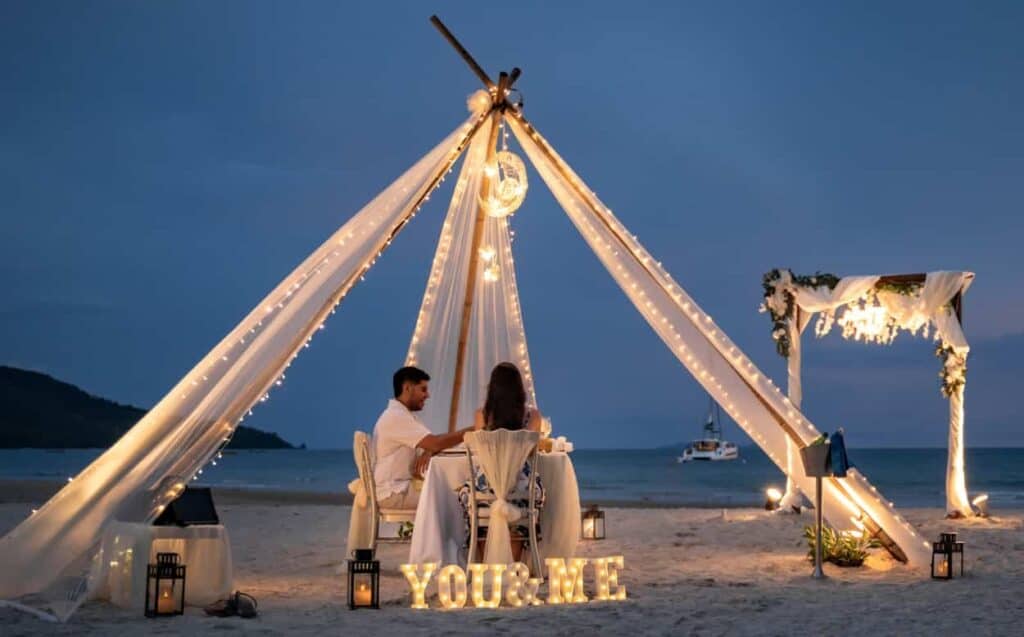 Encontro romântico na praia ao pôr do sol com decoração charmosa e iluminação suave, ideal para casa.
