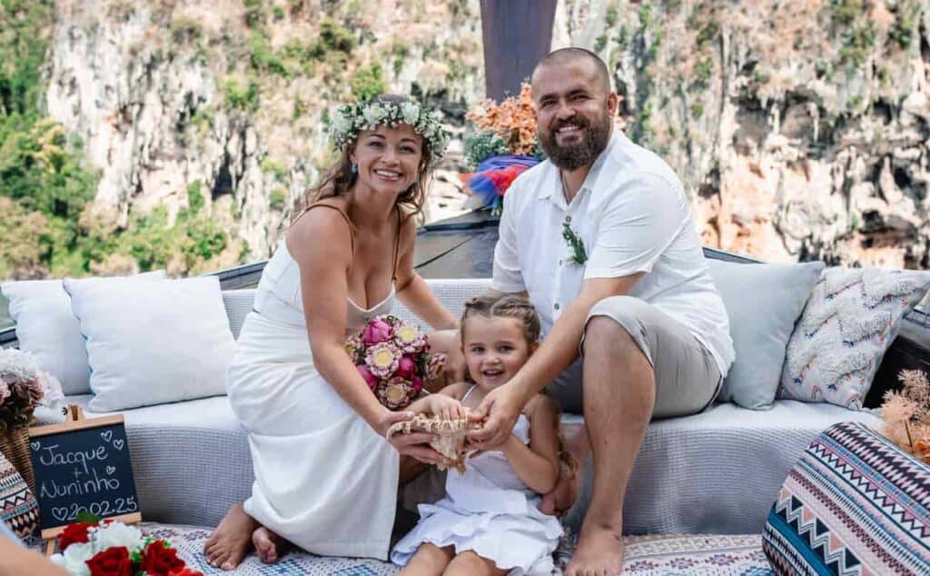 Imagem de um casal e uma criança sorridentes em cerimônia ao ar livre na Tailândia, com cenário natu.
