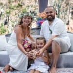 Imagem de um casal e uma criança sorridentes em cerimônia ao ar livre na Tailândia, com cenário natu.